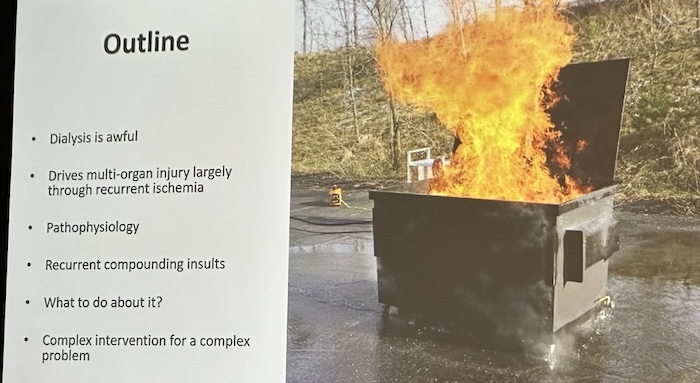 A person standing in front of a screen with a large fire on top of a dumpster Description automatically generated